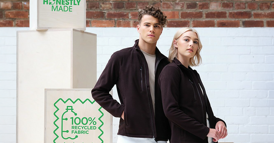 Two models wearing matching black fleece jackets made from 100% recycled fabric, standing in front of eco-branded signage promoting sustainable workwear.