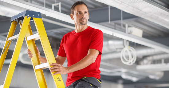 Man in red workwear T-shirt and grey shorts on a ladder inside an industrial workspace, ideal for hot weather. Suitable for embroidery or print branding.