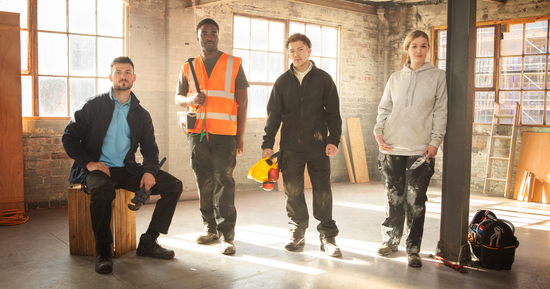 Four construction workers wearing Pro RTX workwear including polo shirts, hoodies, and safety gear, standing in a sunlit industrial workshop with tools and timber in the background.