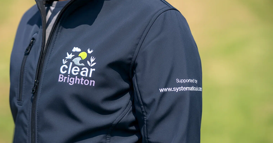 Close-up of a navy softshell jacket featuring detailed embroidered branding for "Clear Brighton" on the chest and a sponsor's website on the sleeve.