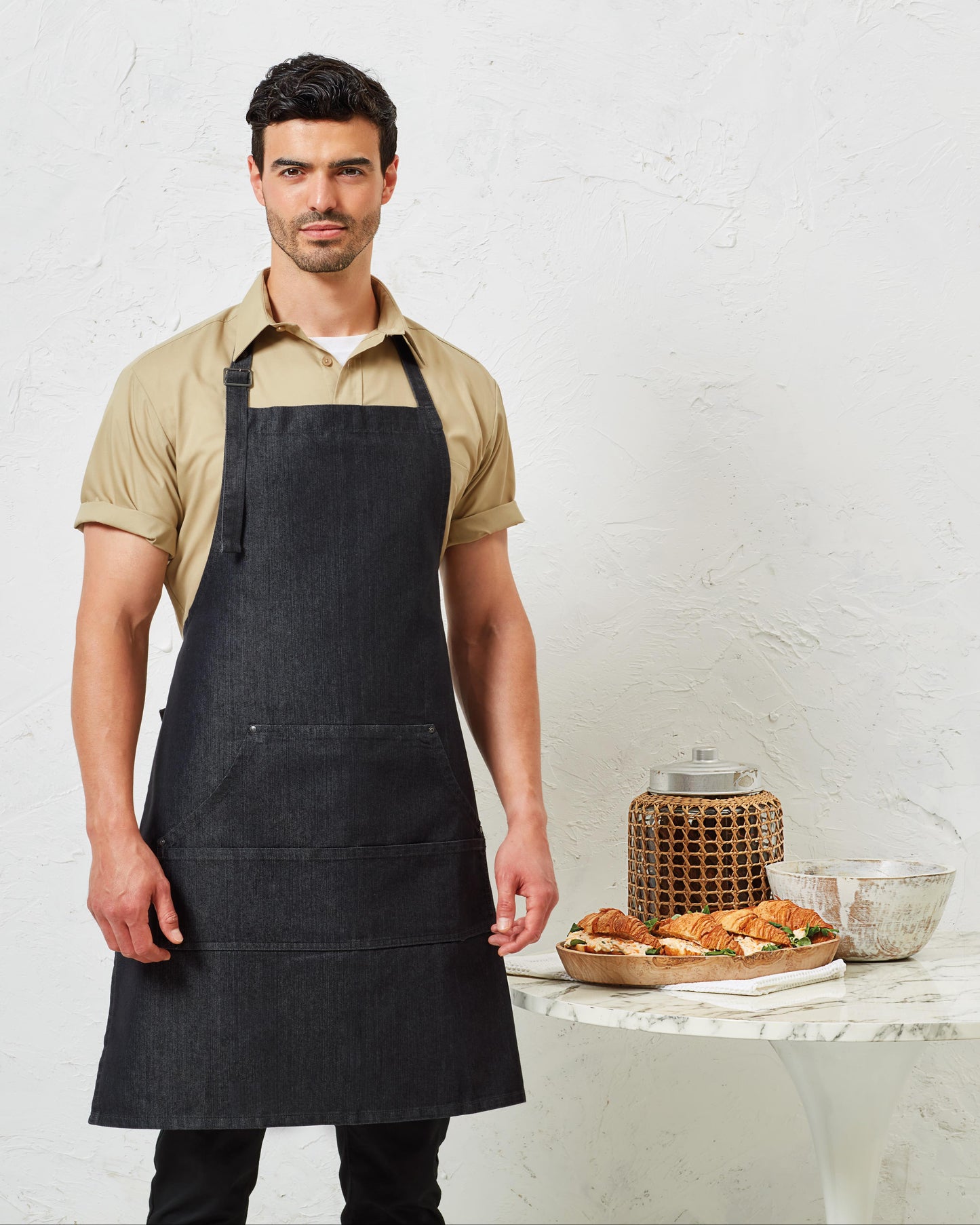 Man wearing a black apron standing in front of a white wall with a table displaying pastries.