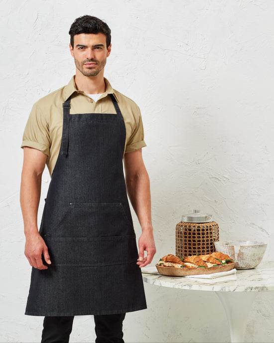 Man wearing a black apron standing in front of a white wall with a table displaying pastries.